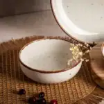 Kuchenne naczynie ceramiczne w kolorze białym z brązową krawędzią, na naturalnym tle, dekoracyjne elementy kwiatowe i owoce, idealne do domowego użytku, nowoczesny design, elegancka estetyka, styl skandynawski, do dekoracji stołu, ekologiczne materiały, ręcznie wykonane, świetne do serwowania i prezentowania potraw, aranżacja wnętrz w stylu boho i minimalistycznym, domowe inspiracje, akcesoria kuchenne.