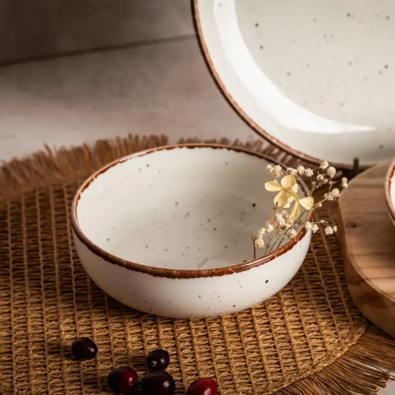 Kuchenne naczynie ceramiczne w kolorze białym z brązową krawędzią, na naturalnym tle, dekoracyjne elementy kwiatowe i owoce, idealne do domowego użytku, nowoczesny design, elegancka estetyka, styl skandynawski, do dekoracji stołu, ekologiczne materiały, ręcznie wykonane, świetne do serwowania i prezentowania potraw, aranżacja wnętrz w stylu boho i minimalistycznym, domowe inspiracje, akcesoria kuchenne.