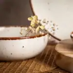 Delikatna biała miska z błyszczącą brązową krawędzią, ozdobiona suszonymi kwiatami i gałązkami, na drewnianej powierzchni, element dekoracyjny domowego zakątka, idealny na naturalne aranżacje wnętrz.
