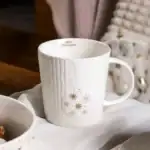 Mleczna świąteczna kubek z zimowym wzorem i napisem Merry Christmas, eleganckie naczynie na zimowe napoje, dekoracyjny element bożonarodzeniowy w domu, ceramiczna filiżanka na święta, dekoracja na świąteczny stół.