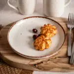 Smażone kotleciki z sera na białym talerzu z czereśniami, rustykalny stół, domowy klimat, ręcznie robione potrawy, elegancka zastawa, naturalne drewno, inspiracje kulinarne, zdrowe jedzenie, komfortowa atmosfera, smak domowego obiadu.