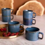 Niebieskie ceramiczne kubki z złotymi paskami na herbatę lub kawę, ustawione na marmurowym blacie z dodatkiem czerwonych malin, idealne do codziennego relaksu i domowego komfortu.