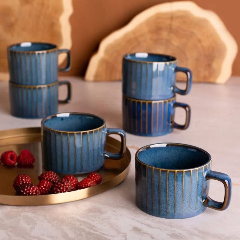 Niebieskie ceramiczne kubki z złotymi paskami na herbatę lub kawę, ustawione na marmurowym blacie z dodatkiem czerwonych malin, idealne do codziennego relaksu i domowego komfortu.
