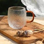 Przezroczysty szklany kubek z pomarańczową rączką na drewnianej desce z makowcami, naturalne światło, rustykalny styl, idealny do napojów, dekoracyjny element domu, wysoka jakość szkła, elegancki i funkcjonalny dodatek kuchenny.