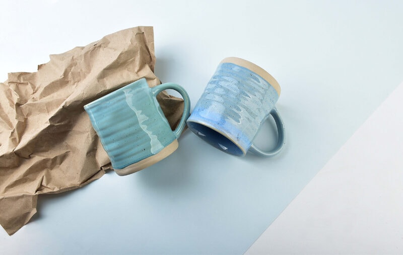 Delikatnie zdobione kubki ceramiczne w odcieniach niebieskiego, ustawione na jasnym tle z kawałkiem brązowego papieru. Idealne do kawy i herbaty, stylowe elementy domu i codziennej atmosfery.