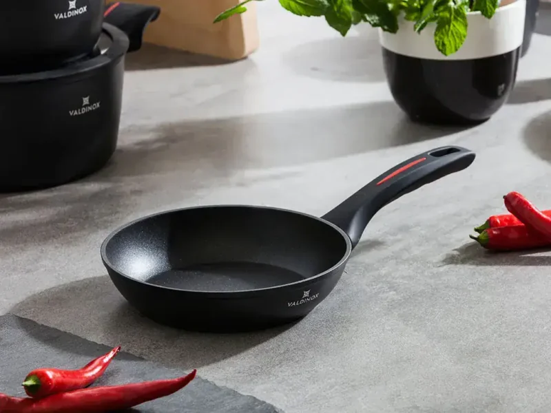Porcelanowa patelnia do smażenia Valdinox na szarym blacie kuchennym z czerwonymi papryczkami i rośliną w czarnym naczyniu, idealna do domowych dań i codziennych posiłków.