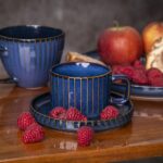 Naczynia ceramiczne w kolorze niebieskim z złotymi prążkami, idealne na kawę lub herbatę, ustawione na drewnianym stole z owocami, takimi jak maliny, jabłka i gruszki, w stylu domowego zacisza.