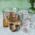 Szklane miarki do odmierzania składników kuchennych na blacie z białej tekstury, na drewnianej tacy, w tle zielona ściana i kwiaty.