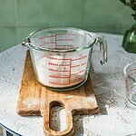 Przezroczysta miarka kuchenna z czerwonymi podziałkami na szklanej powierzchni, umieszczona na drewnianej desce, obok kwiaty w zielonym wazonie, na tle zielonych płytek ceramicznych, idealne do pieczenia i gotowania.