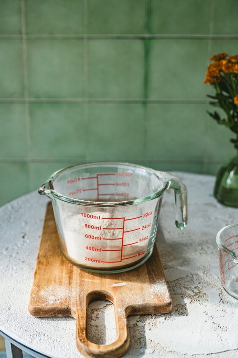 Przezroczysta miarka kuchenna z czerwonymi podziałkami na szklanej powierzchni, umieszczona na drewnianej desce, obok kwiaty w zielonym wazonie, na tle zielonych płytek ceramicznych, idealne do pieczenia i gotowania.