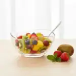 Świeża sałatka owocowa w przezroczystej misie z różnorodnymi owocami, takimi jak kiwi, truskawki, winogrona i mango, na jasnym tle.