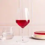 Czerwone wino w kieliszku na stole, obok szklanka wody i kawałek sernika na jasnym tle. Idealne do relaksu i wyjątkowych okazji, wpisujące się w domową atmosferę i smakowe preferencje.