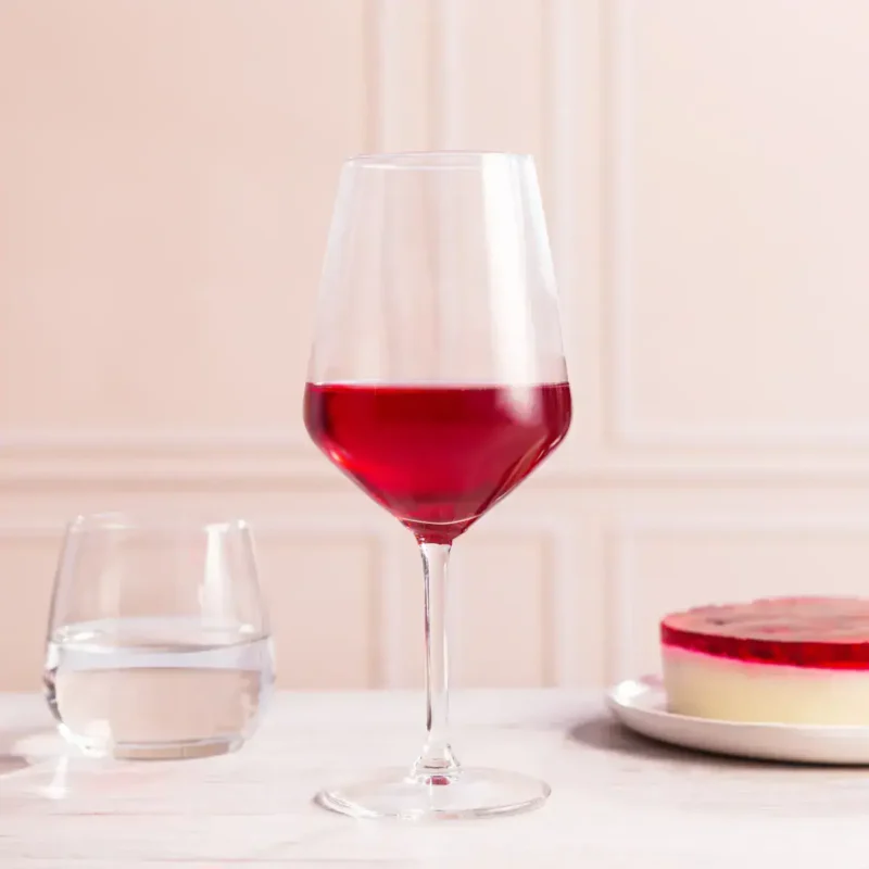 Czerwone wino w kieliszku na stole, obok szklanka wody i kawałek sernika na jasnym tle. Idealne do relaksu i wyjątkowych okazji, wpisujące się w domową atmosferę i smakowe preferencje.
