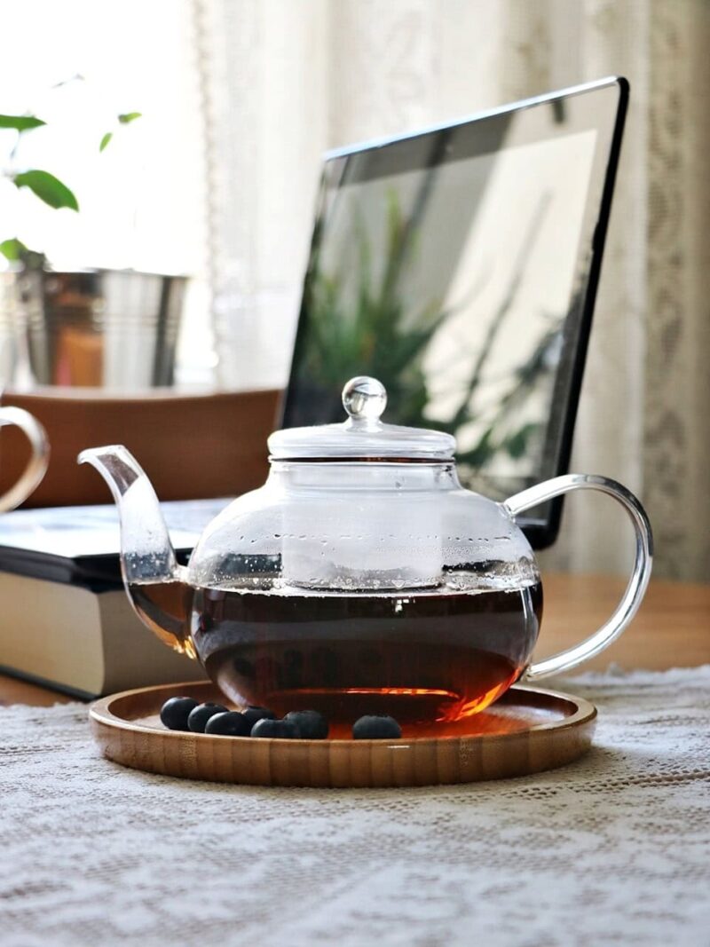 Szklany dzbanek z herbatą na naturalnej tkaninie na tle laptopa, wskazujący na domowe herbatki i relaks w domu.