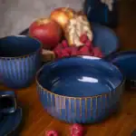 Biala miska ceramiczna z złotymi paseczkami na drewnianym stole z owocami i malinami oraz filiżanki, elegancka zastawa do serwowania owoców i deserów, stylowa i funkcjonalna zastawa domowa, idealna do codziennego użytku i dekoracji stołu, wysokiej jakości ceramika od Domowy Zakątek, wytrzymała i estetyczna oferta naczyń ceramicznych na eleganckie stylizacje i przyjęcia domowe, nowoczesny design i klasyczna kolorystyka dla miłośników eleganckiego wyposażenia kuchni. | Relevance: Tak.