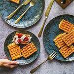 Delikatne gofry na talerzach w bliskim ujęciu, podawane z bitą śmietaną i truskawkami, elegancki deser na śniadanie lub brunch, stylowa zastawa i naturalne światło, tabliczka i czajnik w tle, rustykalny klimat, domowe wypieki, zdrowe i pyszne przekąski, elegancka prezentacja, Pinterest food photography.