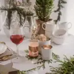 Zimowy stół w stylu bożonarodzeniowym z dekoracjami, świecami, ceramiką i kieliszkiem wina na tle świątecznie udekorowanego domu.