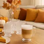 Przytulny narożnik w domowym zaciszu z filiżankami kawy, poduszkami w odcieniach brązu i pomarańczy, dekoracją dyni i jesiennymi gałązkami w szklanej butelce, idealny na jesienne wieczory.