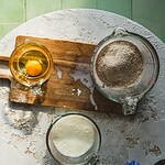 Mieszanka składników do wypieku ciasta, w tym mąka, jajko i mleko, na biały, teksturowany blat, zielone tło w kuchni, z elementami naturalnego światła, podkreślająca domową atmosferę i rękodzieło kuchni.