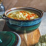 Warzywna zupa w ceramicznym garnku, domowe danie, zdrowa i smaczna, idealna na chłodne dni, w tle kwiaty w wazonie, rustykalny styl kuchni, naturalne składniki, inspiracja do gotowania, kuchnia polska niedroga i pełna smaku, pełnowartościowy posiłek, elegancka zastawa ceramiczna, domowa kuchnia, tradycyjne przepisy, menu na zimę i jesień, odżywcza zupa warzywna, dekoracja stołu, kuchnia w stylu boho, dobrej jakości zastawa, apetyczne danie, zdjęcie ukazujące zdrowe jedzenie, wpisujące się w trendy slow food, domowy sposób na zdrowie, wysokiej jakości ceramika z unikatowym designem, ekologiczne i naturalne składniki, inspiracja do gotowania.