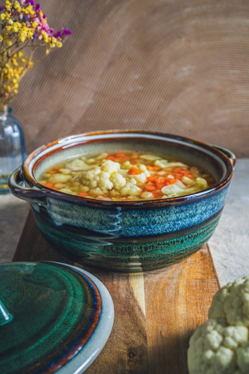 Warzywna zupa w ceramicznym garnku, domowe danie, zdrowa i smaczna, idealna na chłodne dni, w tle kwiaty w wazonie, rustykalny styl kuchni, naturalne składniki, inspiracja do gotowania, kuchnia polska niedroga i pełna smaku, pełnowartościowy posiłek, elegancka zastawa ceramiczna, domowa kuchnia, tradycyjne przepisy, menu na zimę i jesień, odżywcza zupa warzywna, dekoracja stołu, kuchnia w stylu boho, dobrej jakości zastawa, apetyczne danie, zdjęcie ukazujące zdrowe jedzenie, wpisujące się w trendy slow food, domowy sposób na zdrowie, wysokiej jakości ceramika z unikatowym designem, ekologiczne i naturalne składniki, inspiracja do gotowania.
