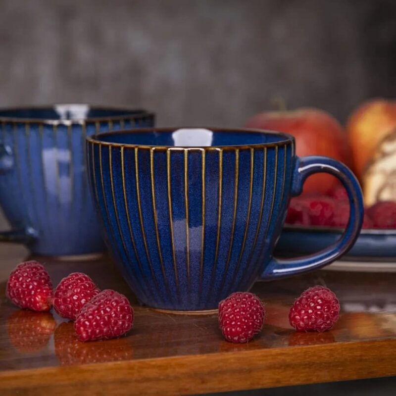 Ceramiczna filiżanka w granatowym kolorze z złotymi paskami, idealna na kawę lub herbatę, na drewnianym stole z malinami i owocami, elegancki element zastawy domowej Domowy Zakątek.