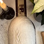 Na zdjęciu znajduje się surowe ciasto do pieczenia chleba z wyrytym wzorem zboża, umieszczone na desce do krojenia, obok świeca w czarnym wazonie i zielona roślina doniczkowa, co tworzy przytulną, rustykalną atmosferę idealną do domowego wypieku chleba.
