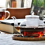 Szklany dzbanek z herbatą na drewnianym talerzu w domowym zaciszu z laptopem i książką w tle. Idealne miejsce do relaksu z aromatycznym napojem w spokojnej atmosferze.