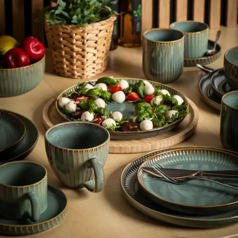 Ceramiczne naczynia i kubki w stylu rustykalnym na stole, w tle kosz z zielonymi ziołami oraz świeże warzywa, idealne do domowego przyjęcia.