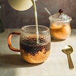 Gęsta śmietanka w szklanej filiżance z miodem, mleko jest wlewane z dzbanka, na stole znajduje się łyżeczka, w tle dekoracja kwiatowa, domowe zwyczaje, inspiracje kulinarne.