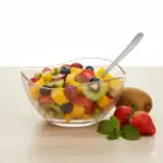 Lekka sałatka owocowa z kiwi, mango, truskawkami i winogronami w przezroczystej misce na jasnym tle.