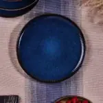 Srebrny talerz ceramiczny w głębi niebieskim odcieniu z dekoracyjnymi złotymi wykończeniami, idealny do eleganckiego stołu, inspirowany stylem Domowego Zakątka, nadaje się na wyjątkowe okazje.